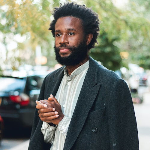 A portrait of a man standing on a Harlem street in autumn. His brown skin is illuminated by the sunlight, giving his face a gentle yet passive expression. His hands are clasped under a charcoal-colored overcoat that rests over his shoulders and his olive-and-white striped shirt.