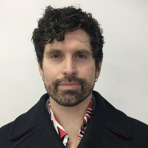 A headshot of a person standing in front of a white wall. They look directly ahead with an expressionless face, perhaps just the hint of a smile. They have dark curly hair and beard stubble. They wear a black peacoat, with a graphic printed shirt peeking out around the collar.