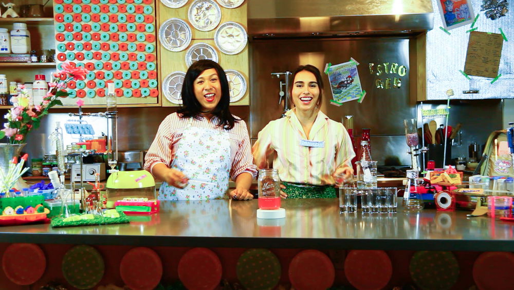 Two transfemme hosts, Maria and Maria, wearing patterned aprons stand posed behind a counter filled with colorful lab equipment, including test tubes, beakers, flasks, shot glasses, and a mason jar filled with a bright pink liquid. The kitchen setting behind them is busily decorated with pink, blue, and silver plates and various posters hanging on the wall, some of which read “Estrogen” and “Estrofem.”