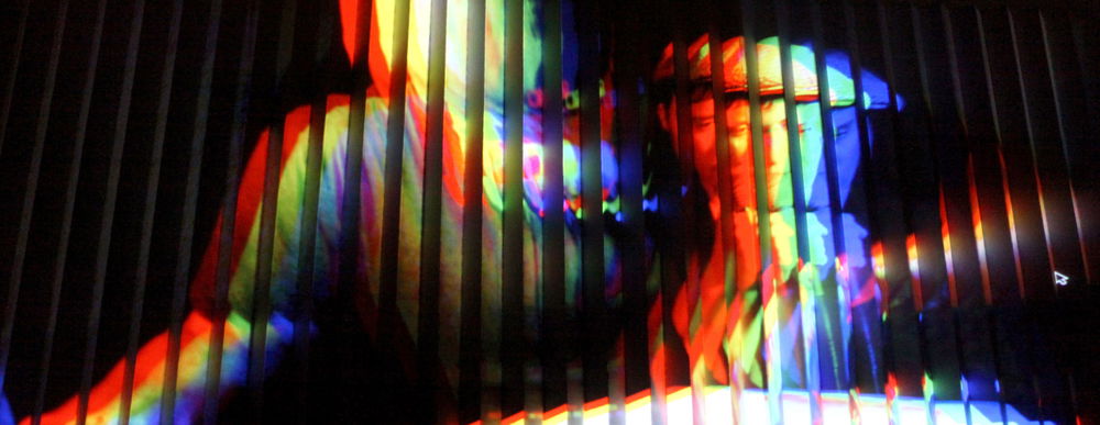 A projection of two obscured figures, one wearing a t-shirt and one wearing a messenger cap, illuminated in red, orange, yellow, green, blue, and purple light. Their image is projected across a number of long vertical slats, multiplying their features across the spectrum of colors, with a black background.