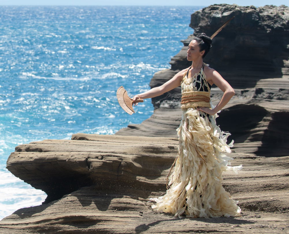 Photo of a woman standing on rocks by the waterfront wearing a netted flowing dress, a heavy cord belt, and holding a plaited fan in her outstretched hand.