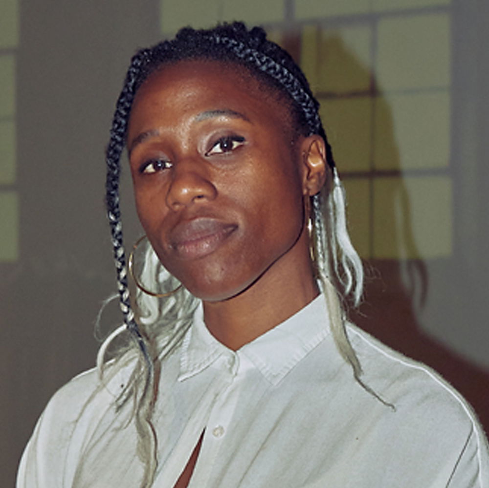 A woman dressed in a white button-down collared shirt poses. Streaks of platinum blonde braids frame her face.