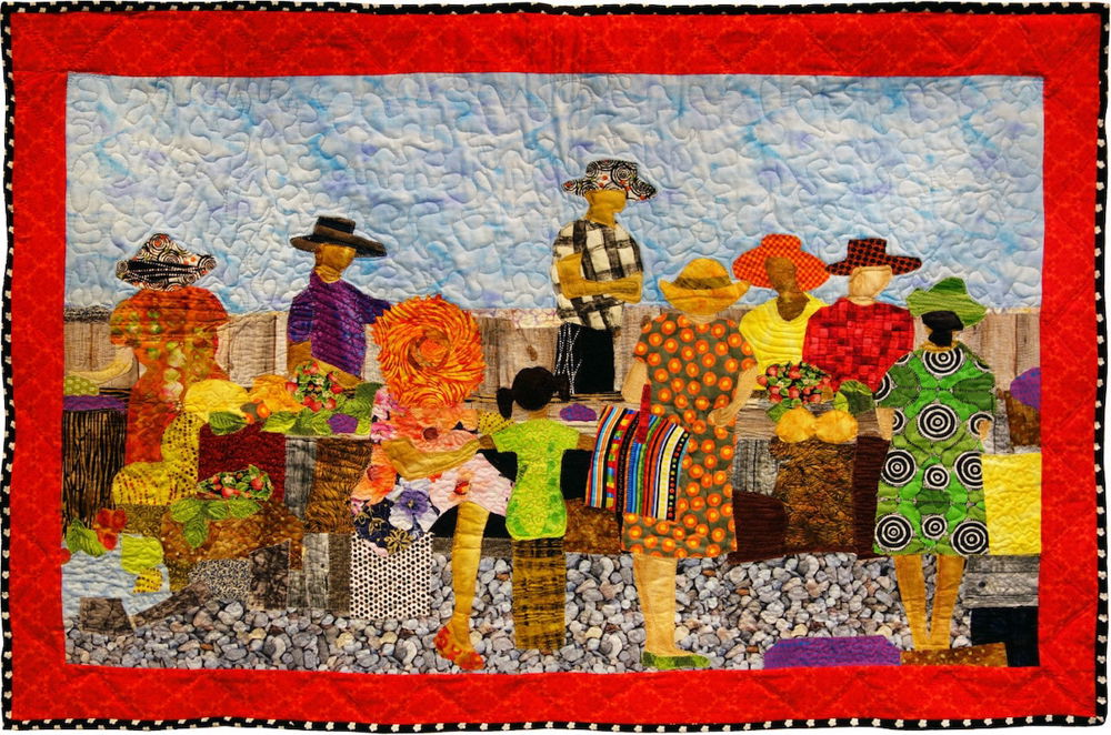 A quilt picturing a vivid scene of nine people standing around a table. Overhead is a light blue sky with a decorative pattern sewn on top. The table is covered in leafy produce.