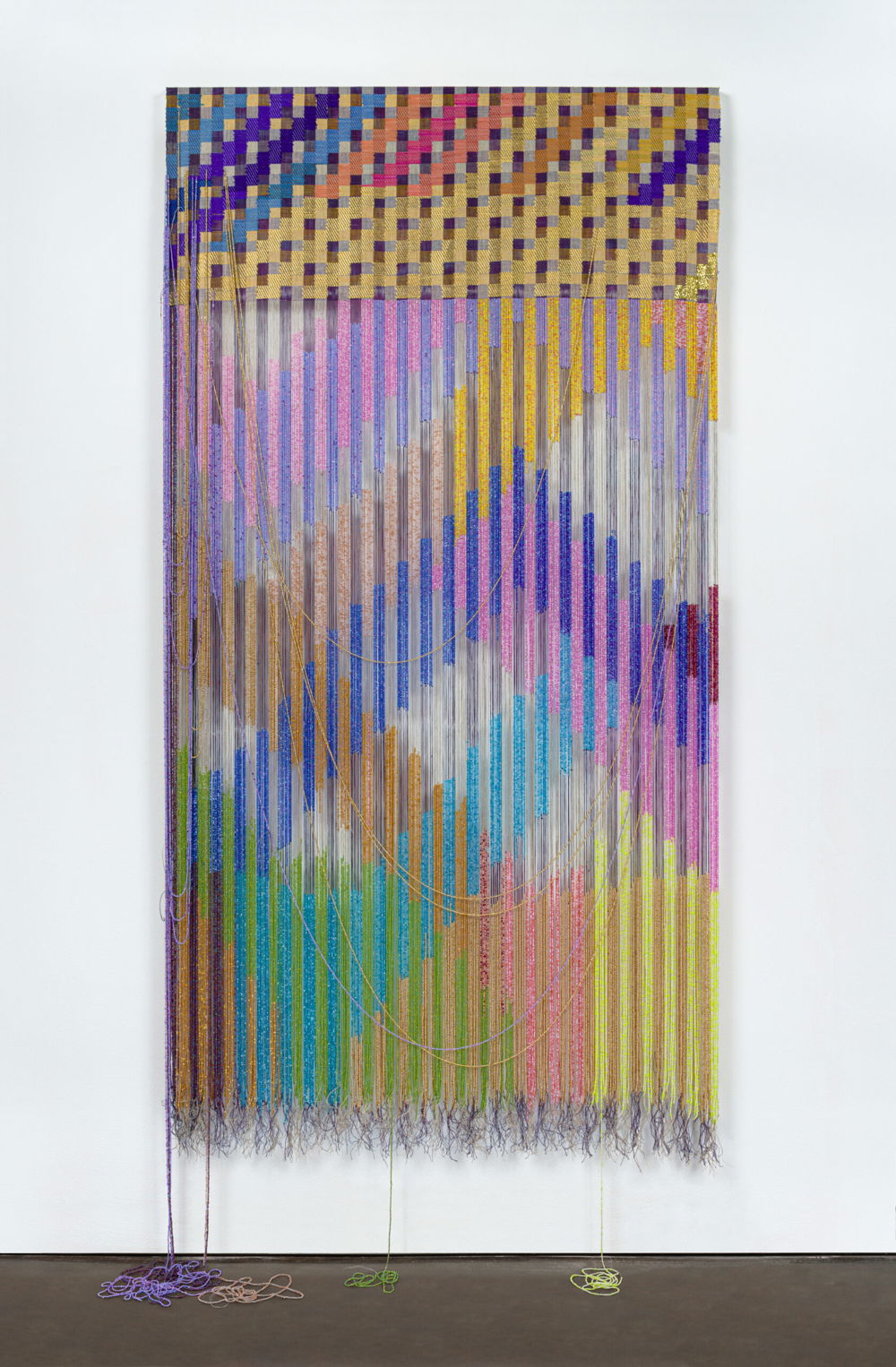A vertical rectangular-shaped tapestry. The top quarter of the composition is a completed tapestry composed of grid pattern in warm purples and taupes overlaid with a stepped diagonal pattern brocaded in light blues, deep purples, warm oranges, pinks, and golds. The bottom three quarters of the composition are unwoven with intricately beaded strands of thread that create a vibrating pattern of intersecting chevrons in yellows, golds, ochres, blues, greens, and purples. A few loose strands are wisped upward creating thin U-shaped lines. Additional loose strands dangle downward to puddle onto the floor.