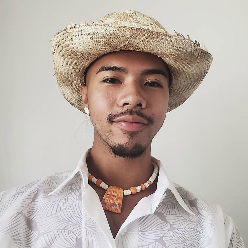 A CHamoru person with light-brown skin and a goatee smiles warmly at the camera. They are wearing a woven pandanus hat, spondylus necklace, and white button-up shirt.