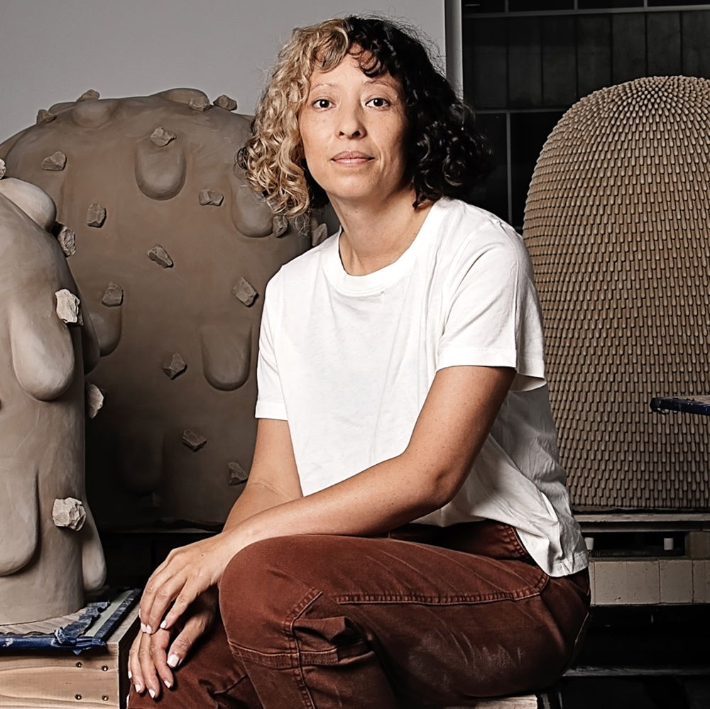 A woman with shoulder-length brown curly hair poses next to a series of clay sculptures as big as her torso. One half of her hair is dyed blonde and she wears a white T-shirt with maroon-colored pants.
