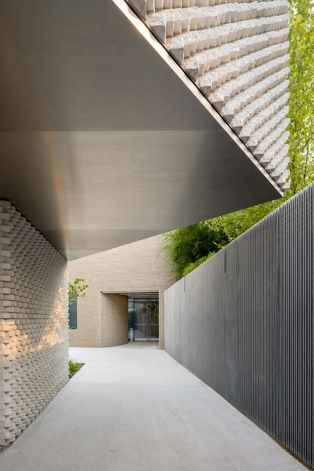 Photo of a view down an outdoor passageway of light, textured concrete, leading to a doorway. A triangular awning hangs overhead.