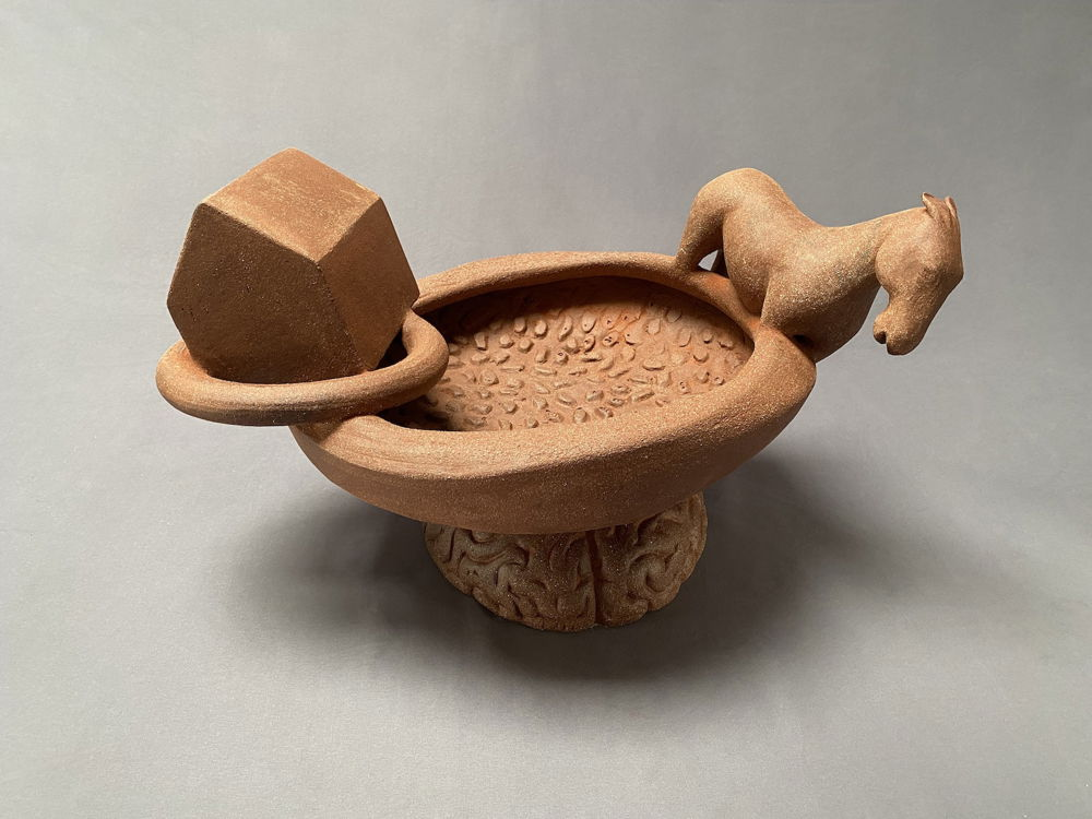 An unglazed red-brown ceramic sculpture of a house and horse sitting on the edge of a bowl, which is balanced on top of a brain