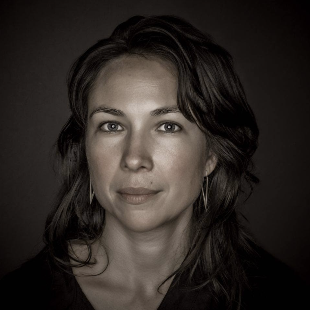 Emily, a Yup’ik woman with long dark hair, wears a black v-neck shirt and pointed earrings against a dark background.