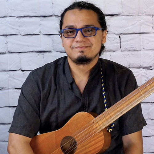 A seated man with his jarana in front of a white brick wall. His dark hair appears pulled back and he gives a passive stare from behind blue framed glasses. His pose is relaxed, his arms hang gently around the jarana, and he gives a very small smile.