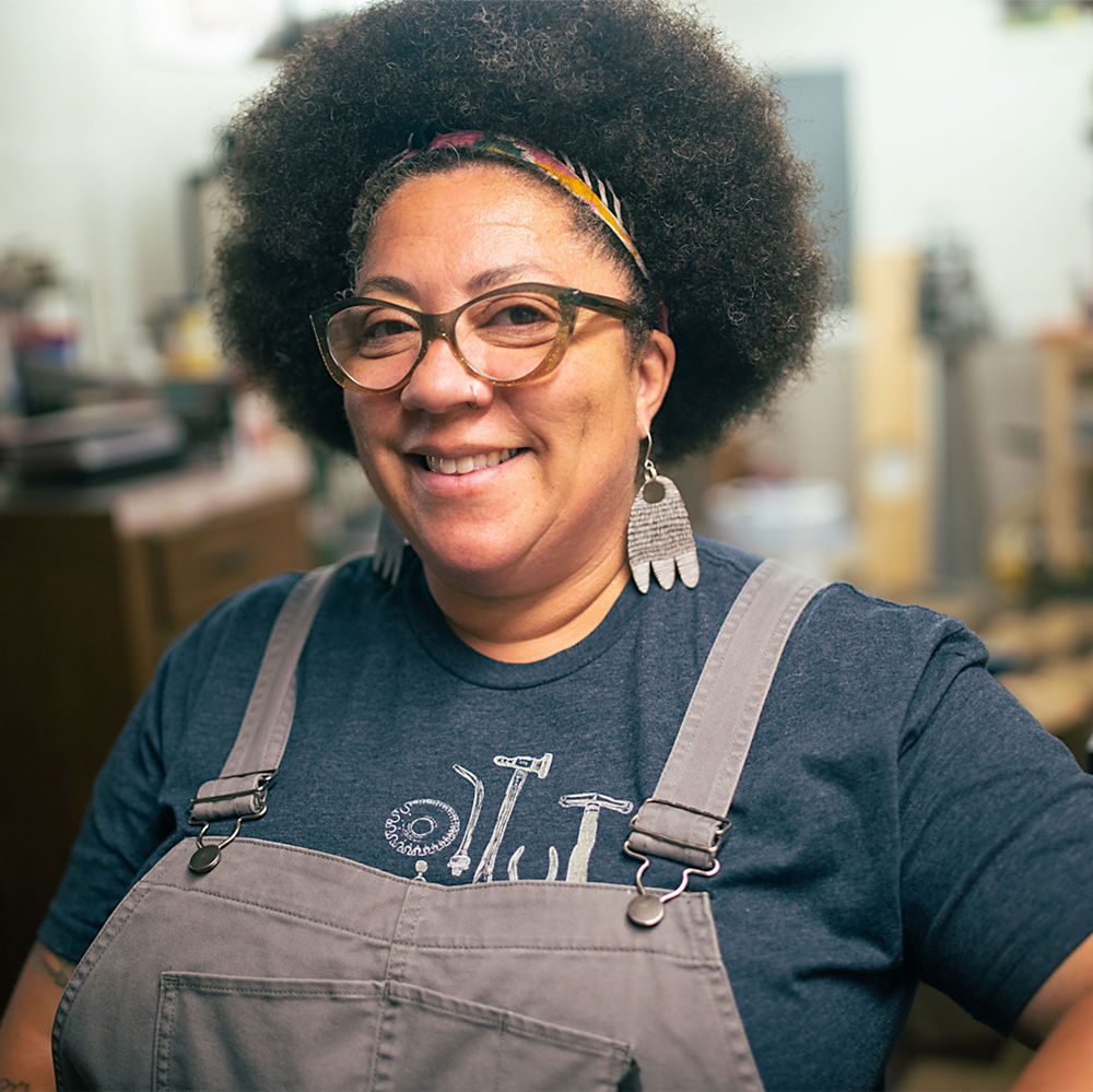 An African American woman smiles at the camera. Her hair is curly and dark in a natural afro style with a colorful scarf tying it back. She is wearing overalls, black-and-white enameled earrings, and sparkly green glasses.