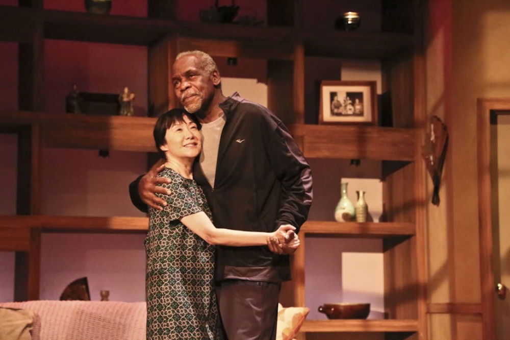 A Black man and a Japanese woman hold hands and embrace on a stage made up to look like a domestic setting