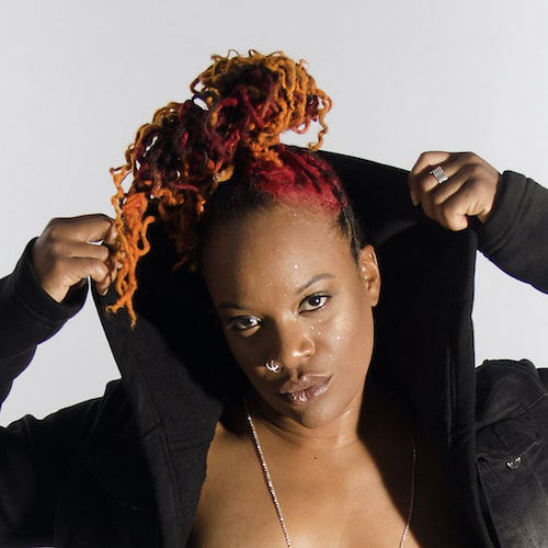 T. Ayo, an African American gender-neutral person with red and orange locks, holds the hood of their black jacket over their head while looking into the camera confidently.