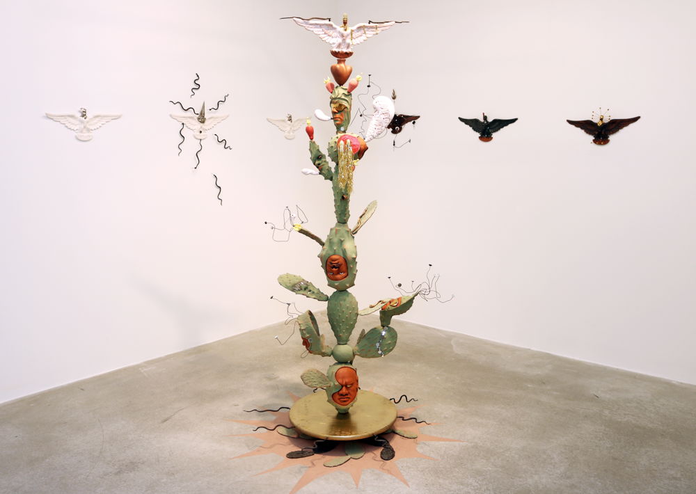 On top of a circular gold base, a totem-like ceramic sculpture depicts different versions of a prickly green cactus and human face stacked on top of each other. At the very top is an eagle with its white wings outstretched. Each wing supports a small snake. On the walls of the gallery behind the sculpture are six additional white and black versions of this eagle and snake.