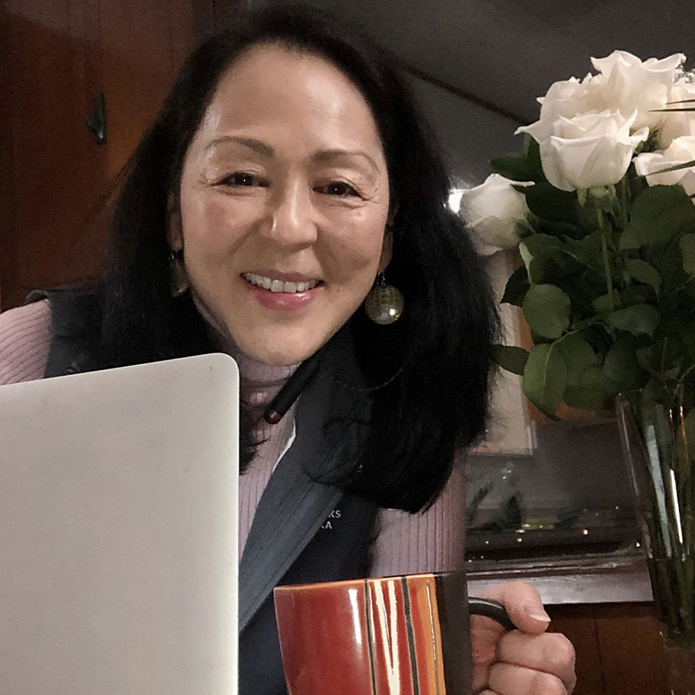 A Japanese American woman with an olive complexion and shoulder-length black hair smiles from behind her laptop computer. She is holding a red coffee cup in one hand, and a vase of lovely white roses rests on the table behind her.