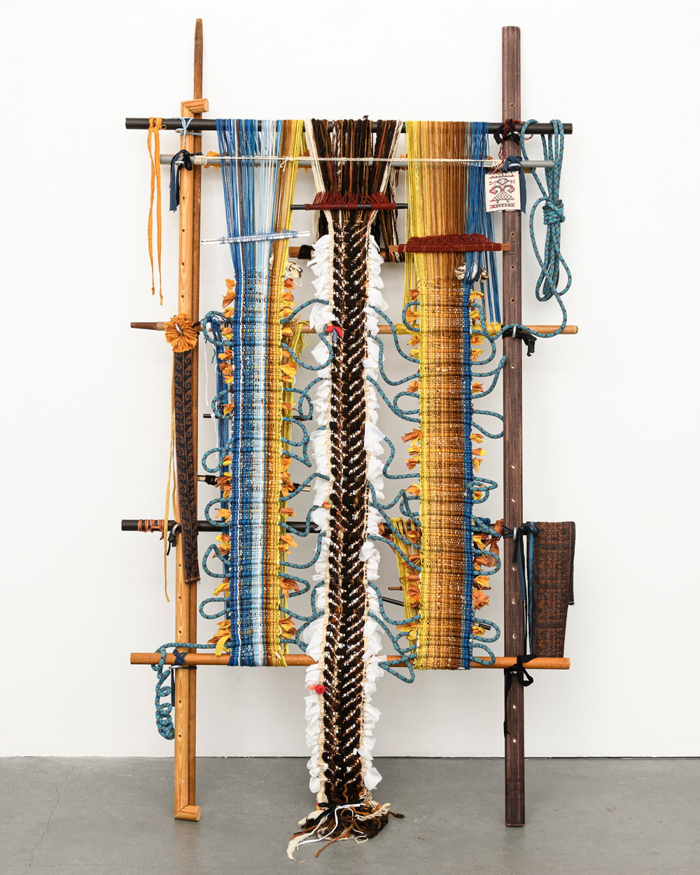 A colorful sculptural weaving that is framed on a wooden loom. There are three vertical panels of finished textiles. The panels to the right and left are gradients of cool blues that transition to warm oranges. The center panel is a black fish-bone pattern on a white background. Waves of blue rope twist between the three panels, connecting them.
