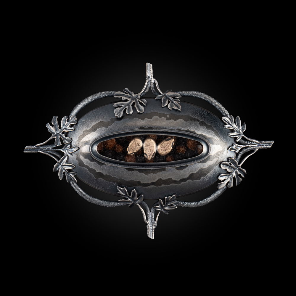 A brooch shaped like a watermelon with vines and leaves along the edges made from sterling silver with horizontal darker bands of nickel silver, photographed on a black background. A central oval opening in the brooch frames three gold seeds in a row on top of balls of human hair.