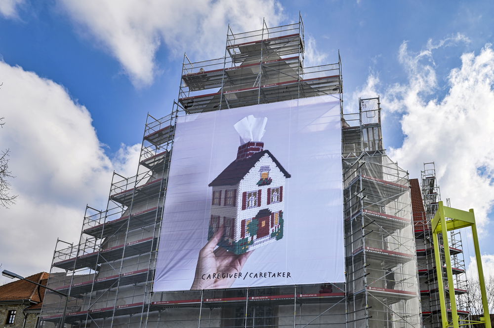 A screen stretched across many floors of scaffolding depicts a picture of a white person's hand holding a tissue box embroidered to look like a little house. The tissues stick out from the house's chimney, like puffs of smoke.