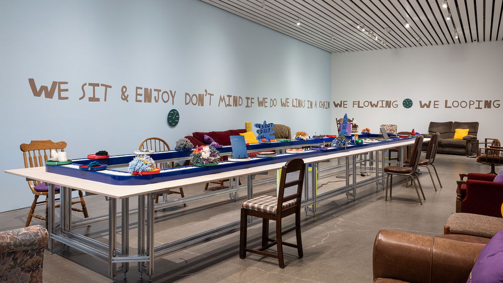 A rectangular conveyor belt rotates colorful and tactile works of art around a rectangular gallery space to different comfortable looking mismatched chairs. Text on the wall reads “We sit and enjoy don't mind if we do we links in a chain we flowing we looping…”