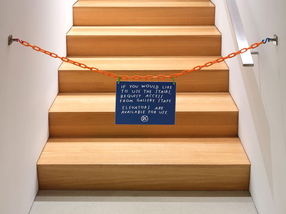 A wooden staircase blocked off by an orange chain and a sign in printed handwriting that reads "If you would like to use the stairs ask gallery staff. Elevators are for use" with an icon of a crossed-out staircase.