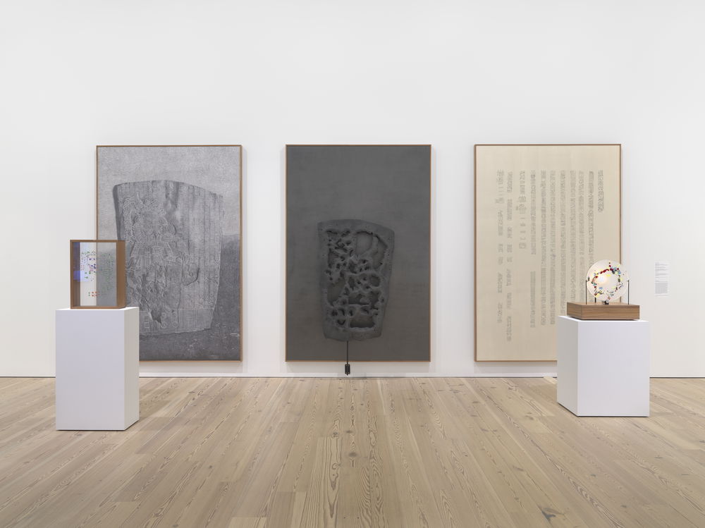 Installation view of three wall-mounted panels and two small sculptures set on pedestals in front of them, all referencing La Mojarra Stella, a mesoamerican engraved artifact. The first panel is a realistic graphite drawing of La Mojarra Stella. Propped in front of it is a transparent film printed with colored markings matching the artifact itself, like a map key. The second panel is an abstract version of the artifact mounted on a dark background with holes as if parts have been cut out. The third drawing transcribes the glyphs carved on the stone. The second sculpture is a white circle with colorful markings on its surface.