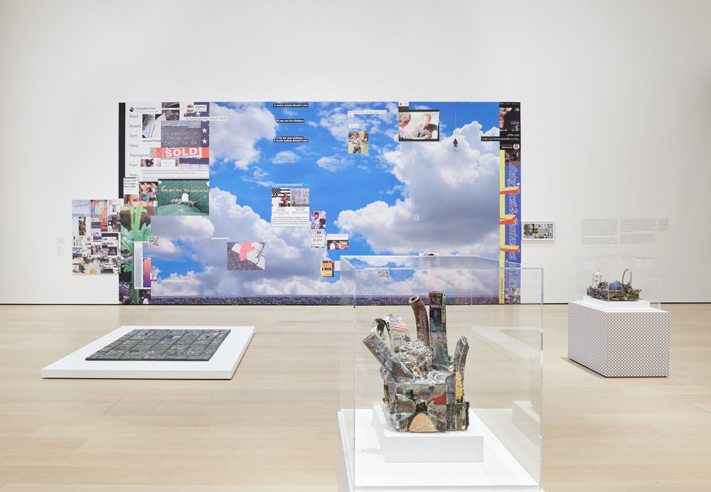 An installation shot in a white gallery space. On the back wall, directly facing us, is an enormous mixed-media painting installed with the bottom of the canvas resting on the floor. The horizontally-oriented work features a crisp blue sky filled with scattered clouds. Superimposed on top of the sky are scattered boxes of images and text that resemble pop-up windows on a computer screen. A few of the "pop-up windows" float off the canvas and onto the white wall.