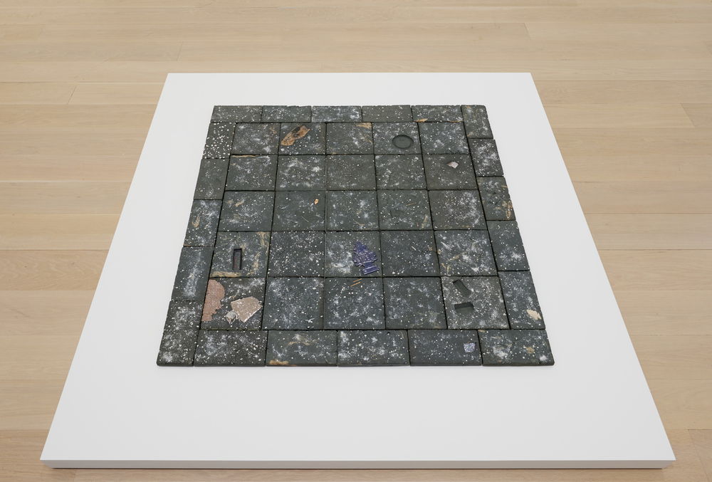 A flat rectangle-shaped sculpture lies flush on a shallow white platform, raised only a few inches from the floor. The sculpture resembles a patch of filthy gray stone tiles splattered with dirt, garbage, and bird droppings.