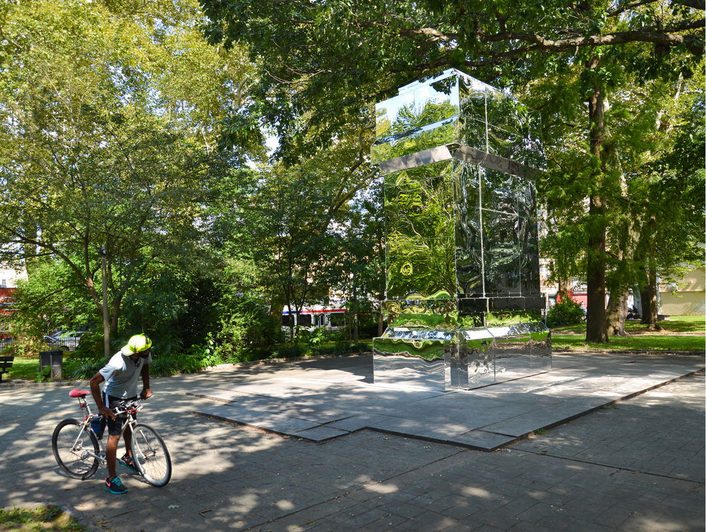 A Black man halts his bike ride to check out a column made of mirrors erected in a green space. The sturdy rectangular column reflecting the surrounding foliage is not immediately perceptible.