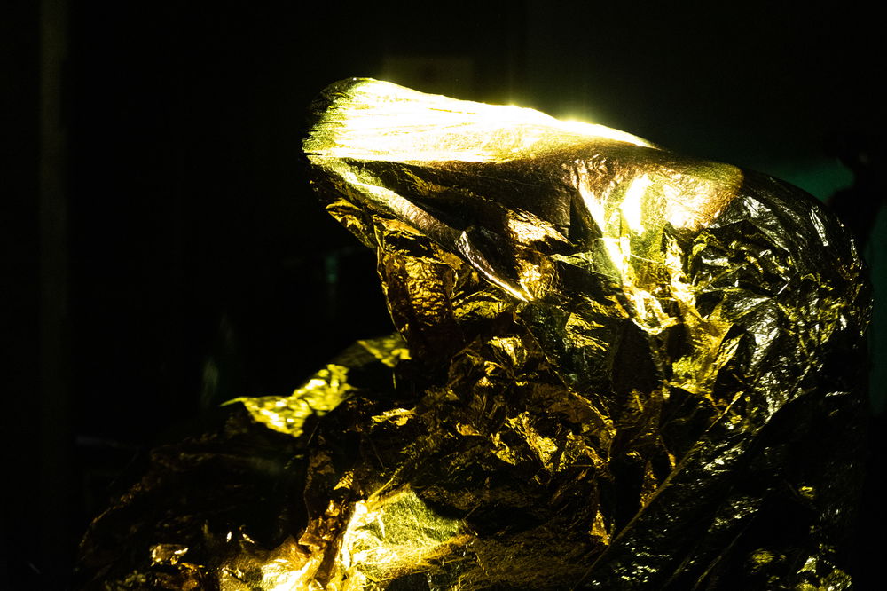 A dark photograph with a bundle of gleaming shiny gold fabric illuminated by an unseen light source.