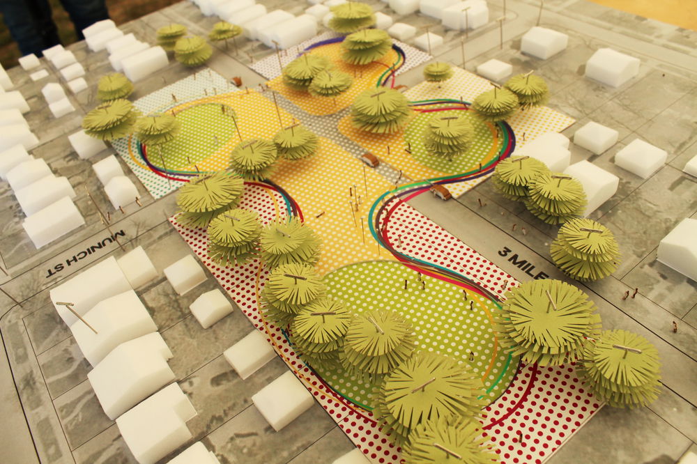 A close up of a model of a greenspace situated between blocks of city buildings. The green space is indicated by curvy organic colorful lines swirling around a yellow background and dotted with green circular fringe trees.