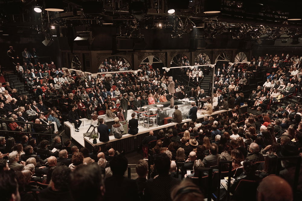 A wide shot of a narrow horizontally-oriented stage situated in the center of a theater surrounded by stadium style seating. Actors sit around the stage surrounding a suited actor standing on metal counters.