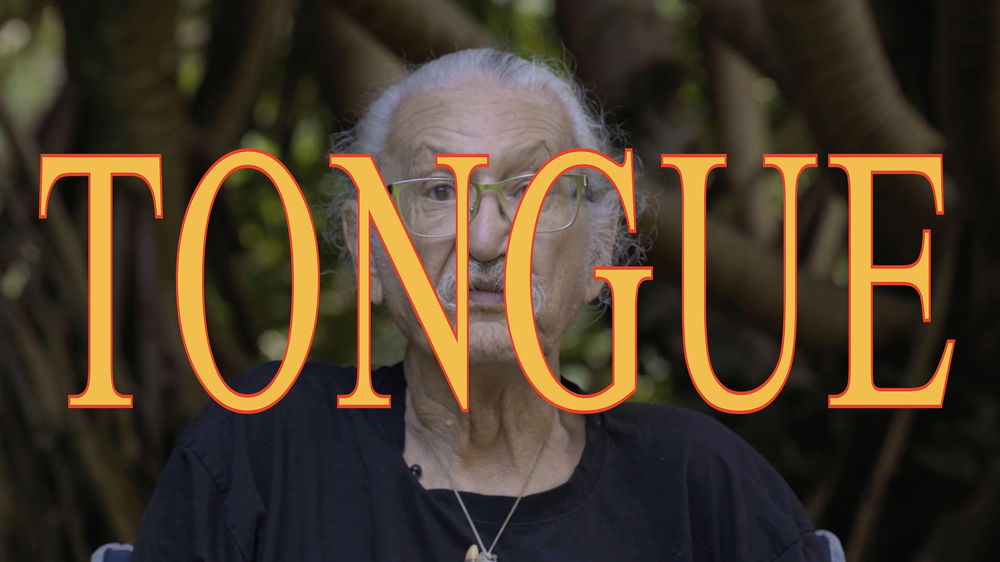 A close-up of an older adult man's face. He has a light-brown complexion and long white hair tied back behind his head. He wears silver-rimmed glasses and a plain black t-shirt. He raises his eyebrows slightly, but otherwise has a neutral expression. The word “Tongue” is written across the image, partially obscuring the man's face.