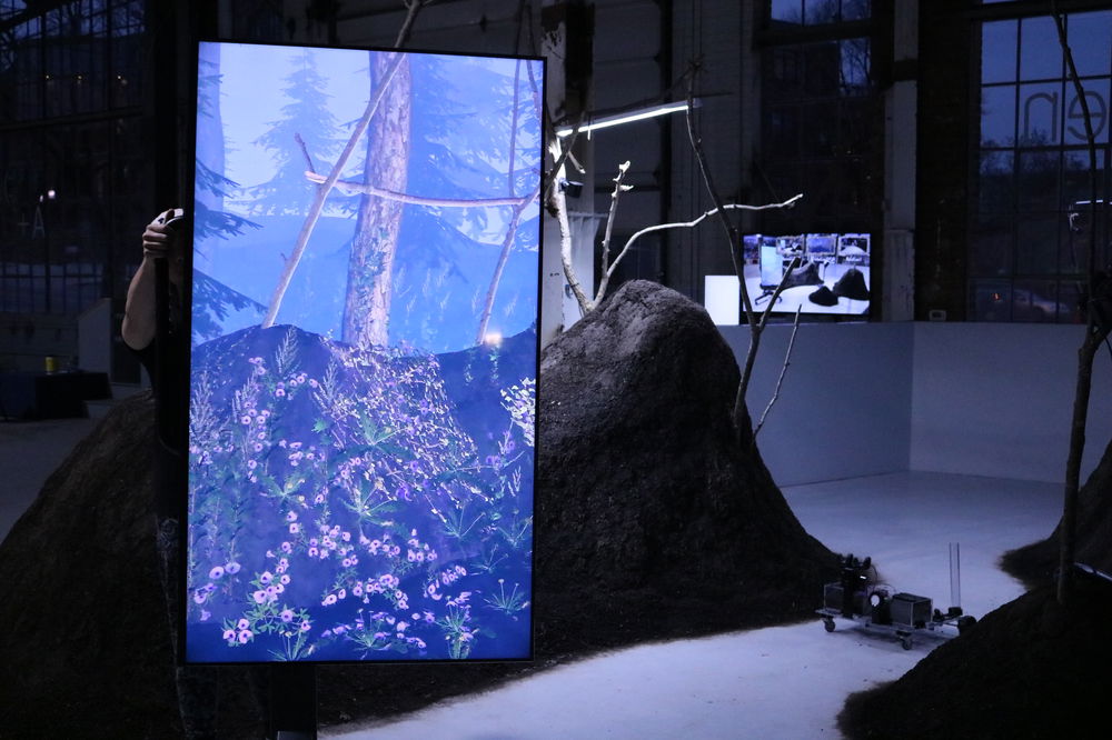 A vertically-oriented screen portrays an eerie image of a blue tinted forest whose ground is covered with purple wildflowers. Just behind the screen is a mound of dirt with tree branches sticking out.