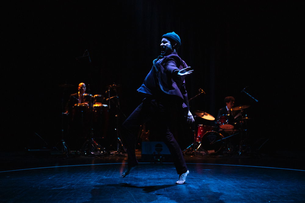 A performer, seen in profile, on a dark stage as they skip-jump to our right. They hold their arms out at their sides with their palms up and scream. Two musicians seated at drum sets are dimly visible behind the performer.