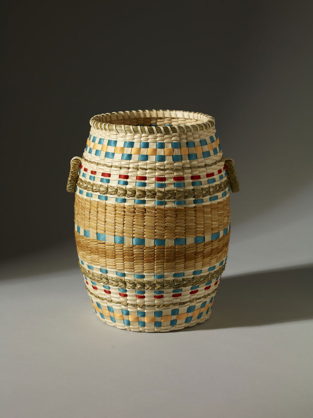 A beige woven basket dotted with teal and red fibers. A handle hangs down from each side.