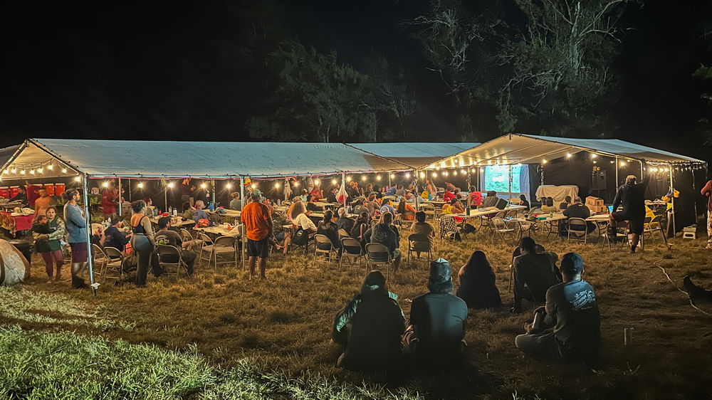 People gather to watch a film screening outdoors under a white tent in the evening.