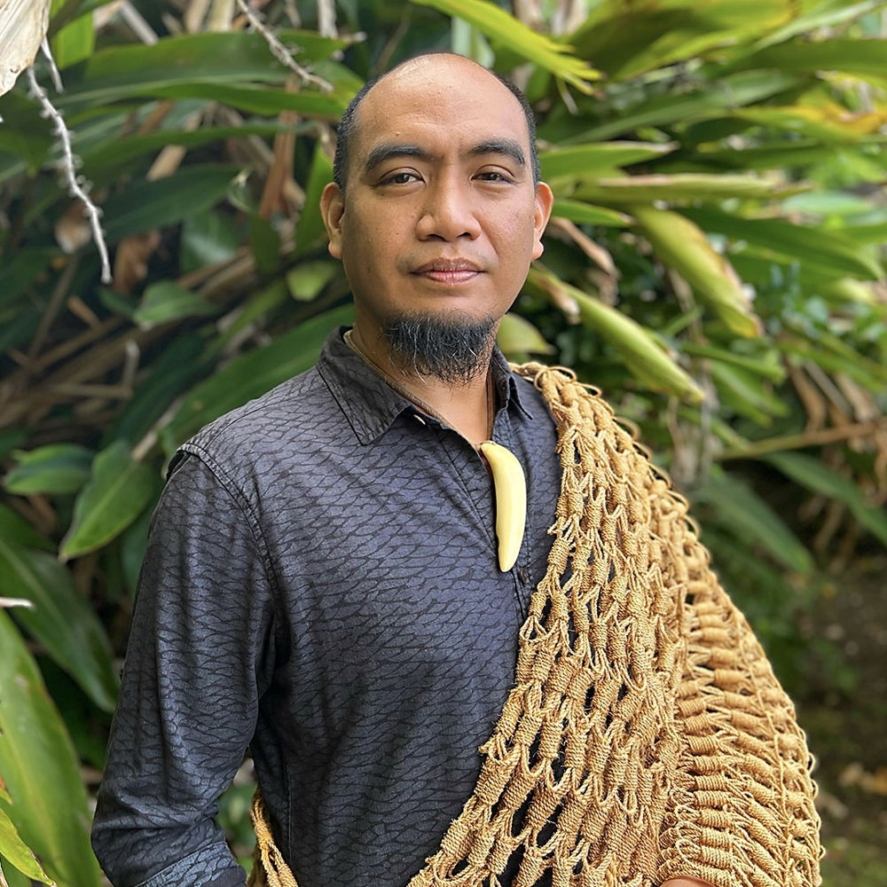 A man with brown skin and black goatee poses wearing a black, long-sleeved collared shirt with a net woven from natural fibers draped over his left shoulder and a carved pendant hanging from his neck. Behind him is a large bush with elongated green leaves.