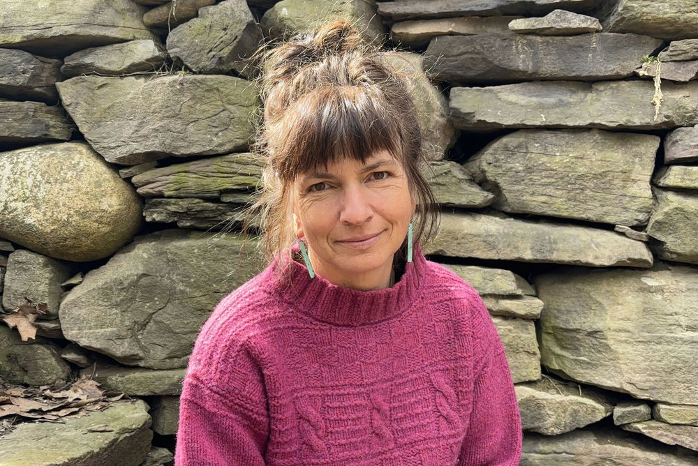 Thea, a woman with brown hair swept up into a messy bun and wearing a bright pink sweater, sits on stone steps beside a towering stone wall.