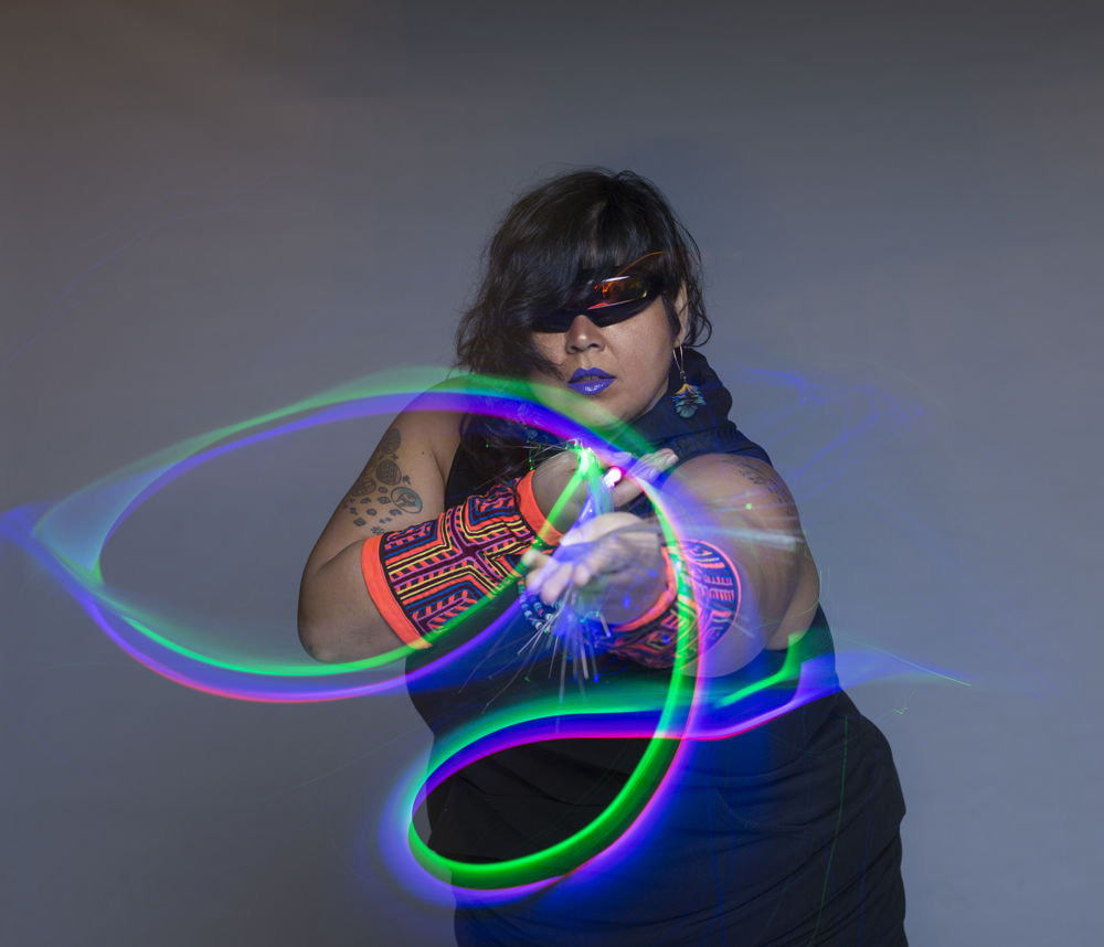 Mother Cyborg, a Latin woman with cyclops glasses and neon armbands on is surrounded by glowing streams of colorful light that she seems to be manipulating.