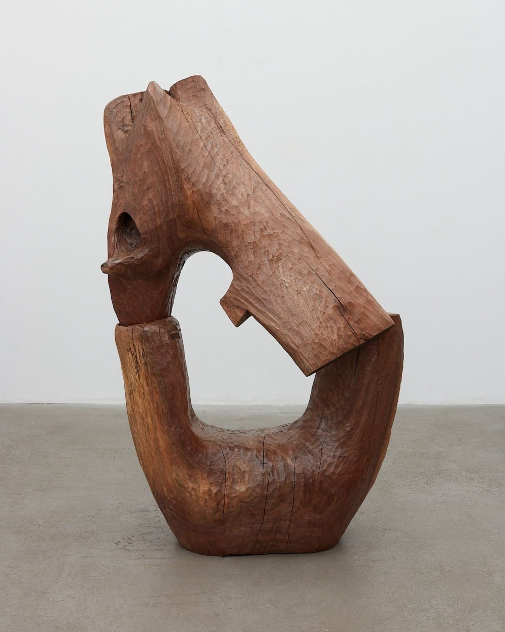 Photo of a wooden sculpture formed of two curved pieces of wood resting together in a keyhole shape. Carving marks and woodgrain are visible on the surface.