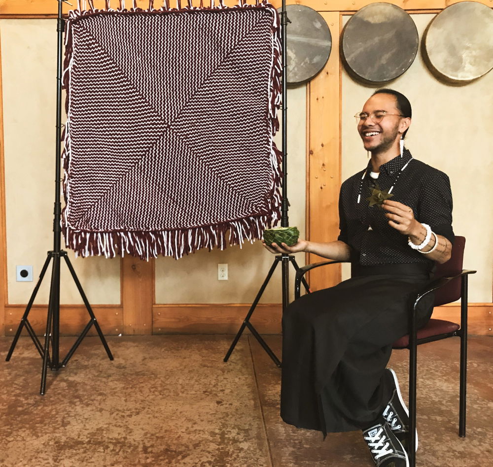 The artist poses next to a maroon-and-white blanket with fringe hung in a metal frame. The points of four triangles meet in the center to form a big X. The pattern creates a zigzag of woven lines moving from the edge of each triangle to the center.