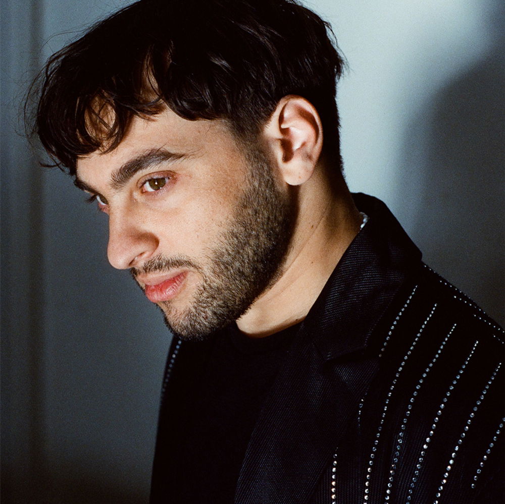 A mixed-race person with tan skin, mid-short cropped hair, short beard, and a black-and-silver outfit gazes beyond the camera.