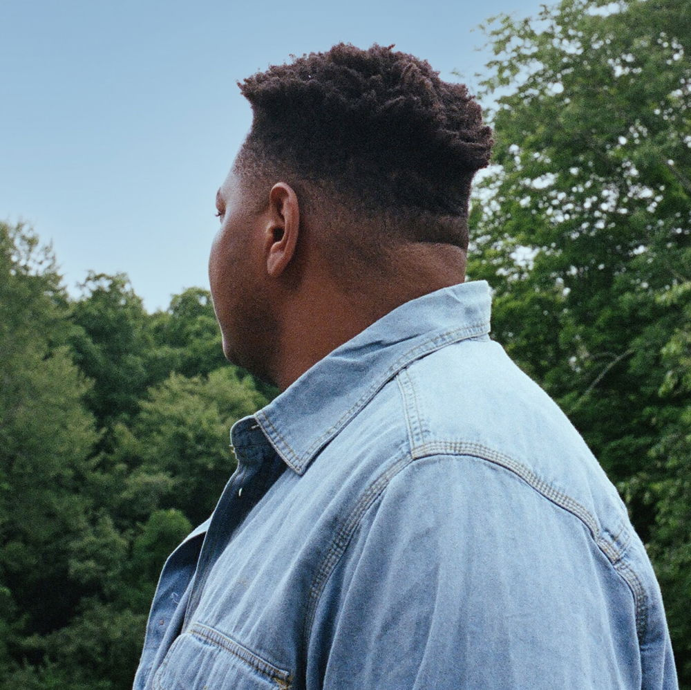 Darius, a Black man with cropped hair, wearing a blue-jean shirt, stands contemplatively upon a balcony in profile surrounded by plush green trees, looking out into the distance.