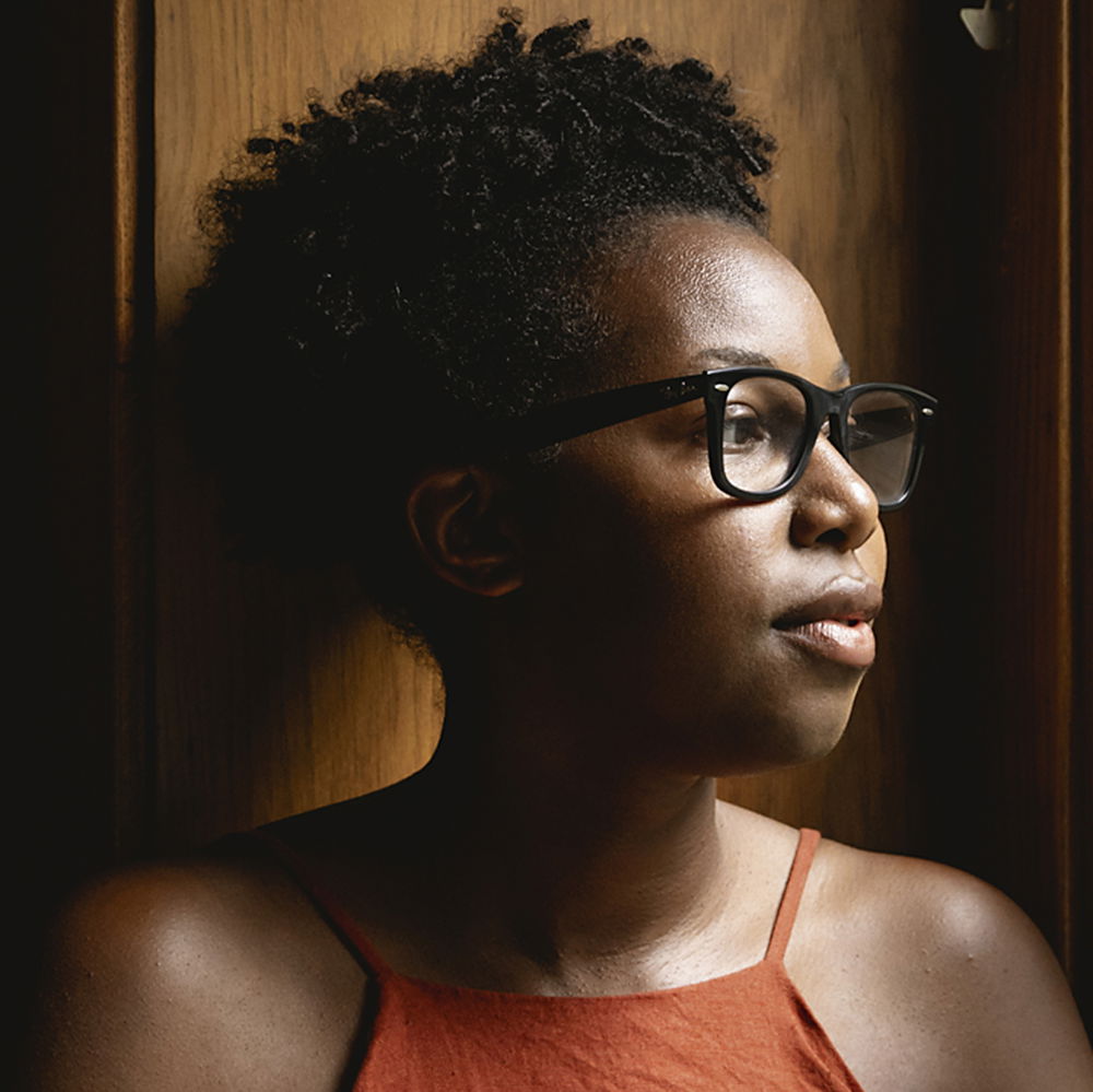A dark-skinned woman wearing a red sleeveless top and black-rimmed glasses looks out to her left.