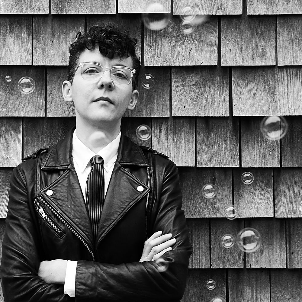 A white, transmasculine, nonbinary person with short, dark hair stands against a shingled wall. Their arms are crossed, and they wear a white shirt, black-and-white tie, and black leather jacket. Bubbles float through the picture; one hovers over their Adam’s apple.