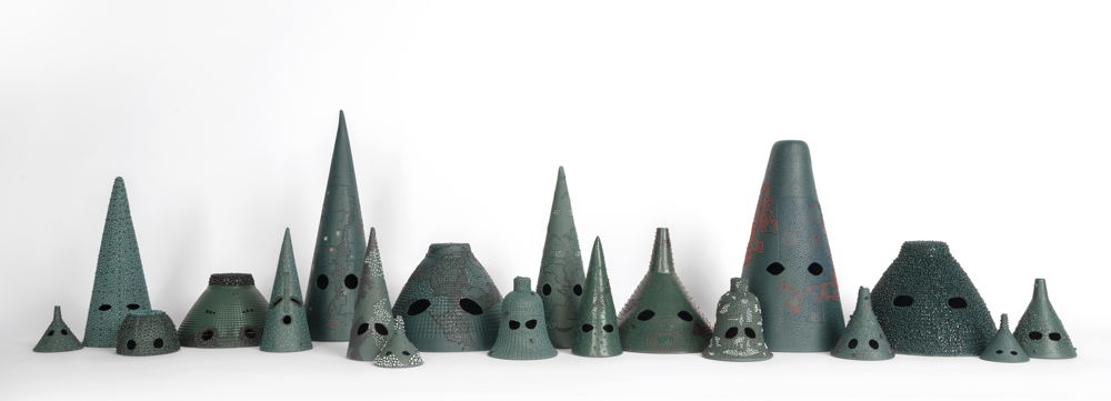 A row of sage-green ceramic sculptures lined up in front of a white backdrop. The sculptures vary in height but are all conical, spherical, or cylindrical in shape. Near the base of every sculpture are two eye-shaped cutouts.