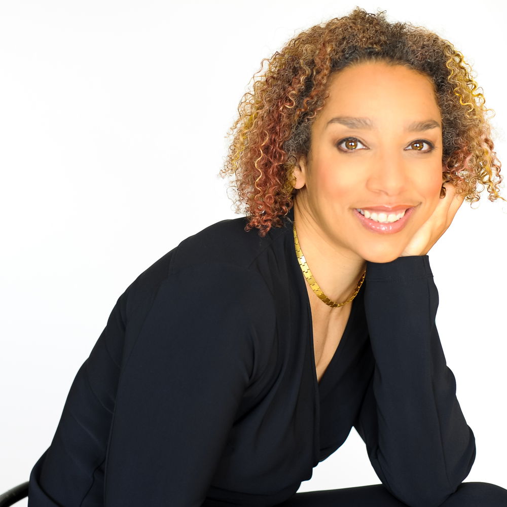 Alice, a Black woman, smiles brightly with her head in her hand. She has light brown skin, curly shoulder-length hair with bronze highlights, and wears a black blouse and sleek gold necklace.