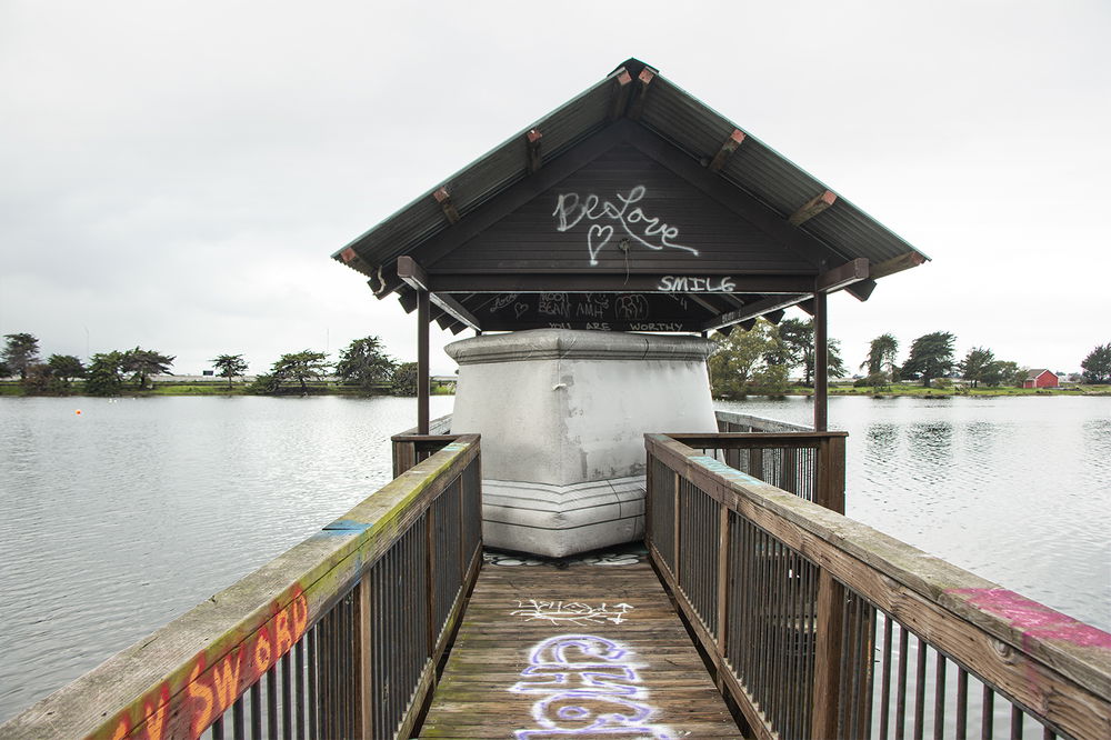 At the far, covered end of a wooden pier sits an inflatable monument base, with no statue sitting upon it, squeezed at an angle to fit into the space. The surface of the inflatable has a concrete pattern that appears to have been painted over, as if layers of protest graffiti have been whitewashed.