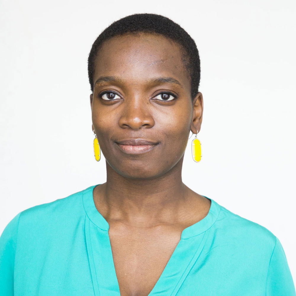 Njideka, a Black woman with short dark hair, stands against a white background. She wears an emerald green top, yellow dangling earrings and smiles slightly at the camera.