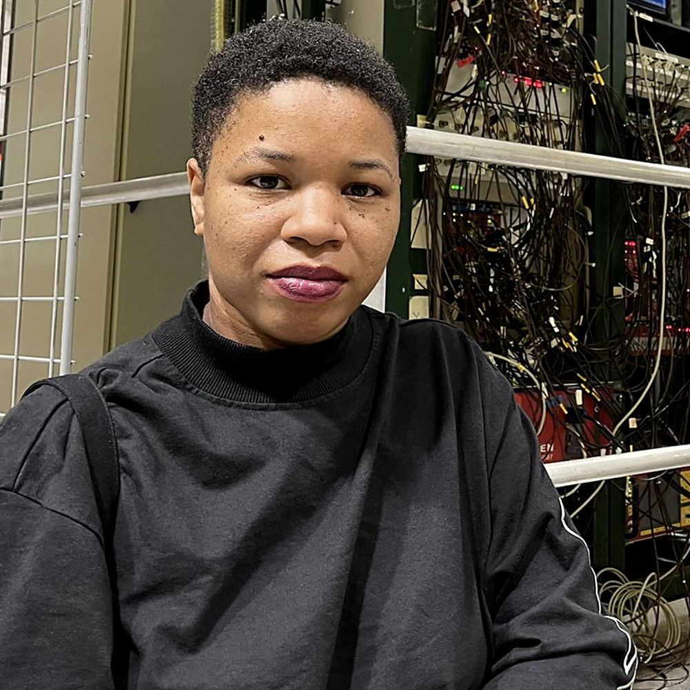 A Black person with brown skin, short hair, and red lipstick standing inside of a large warehouse in the GBAR Experiment at CERN, the largest particle physics laboratory in the world.